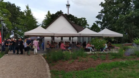 Ein modernes Gebäude mit einem weißen Dach und überdachten Terrassen. Viele Menschen sitzen draußen und genießen die Umgebung in einem Park.