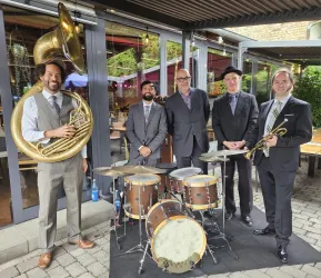 Eine Musikgruppe steht vor einem Restaurant. Sie tragen Anzüge und spielen verschiedene Instrumente wie Tuba, Trompete und Schlagzeug.