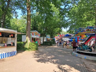 Eine belebte Festplatz mit bunten Fahrgeschäften und Ständen. Dicke Bäume spenden Schatten und schaffen eine fröhliche Atmosphäre.