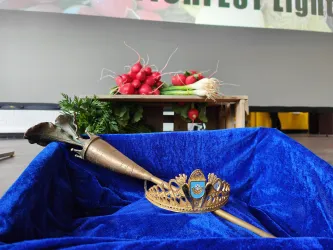 Eine glänzende Krone und ein dekorierter Stab liegen auf einem blauen Tuch. Im Hintergrund sind frische Radieschen und Lauch zu sehen.
