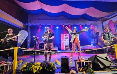 Eine Band spielt live auf einer Bühne mit bunten Lichtern. Der Sänger hat die Arme in die Luft gehoben und die Atmosphäre ist festlich.