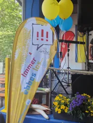 Ein Banner mit der Aufschrift "Immer eppes los" steht auf der Bühne. Im Hintergrund sind bunte Luftballons und Blumenarrangements zu sehen.