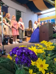 Eine Blumenpracht mit lila und gelben Blumen im Vordergrund. Im Hintergrund stehen mehrere Personen auf einer Bühne und hören einer Ansprache zu.