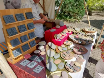 Ein Verkaufsstand mit vielen handgefertigten Steinobjekten und Dekorationen. Im Hintergrund steht eine Person, die an einem weiteren Stein arbeitet.