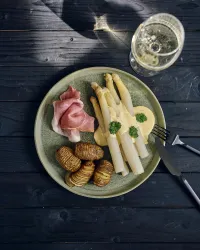 Ein schön angerichteter Teller mit Spargel, Schinken und spiralförmig geschnittenen Kartoffeln. Im Hintergrund steht ein Glaspfang, das ein Getränk enthält.