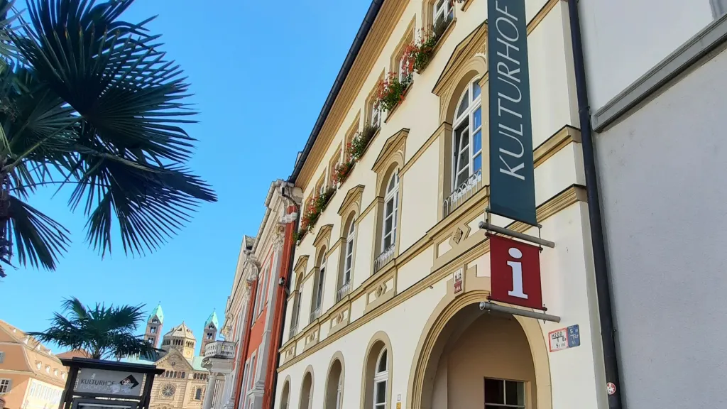 Die Außenfassade der Tourist Information in Speyer mit einer Palme und dem Dom im Hintergrund