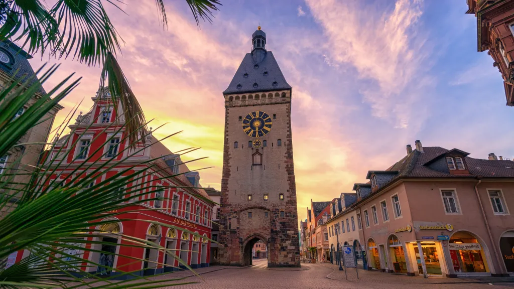 Vorplatz mit Palme, Gebäuden und Stadttorturm bei Abendstimmung