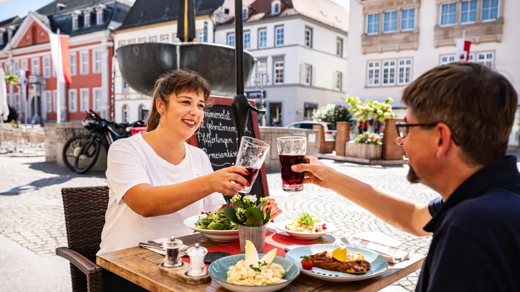 Zwei Personen sitzen im freien an einem Tisch gedeckt mit Essen und stoßen mit gefüllten Gläsern an