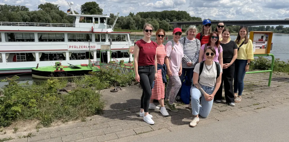 Eine Gruppe Menschen steht am Rheinufer vor einem Schiff
