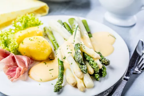 Ein weißer Teller mit Kartoffeln, Spargel und einer Scheibe Schinken, darüber eine helle Soße