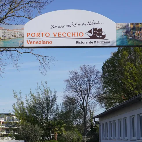Ein Schild mit der Aufschrift "PORTO VECCHIO Veneziano Ristorante & Pizzeria". Es steht in einer grünen Umgebung mit Bäumen im Hintergrund.