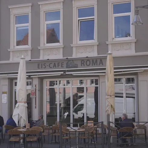 Ein charmantes Eiscafé mit der Aufschrift "EIS-CAFÉ ROMA". Vor dem Café sitzen Gäste an Tischen im Freien.