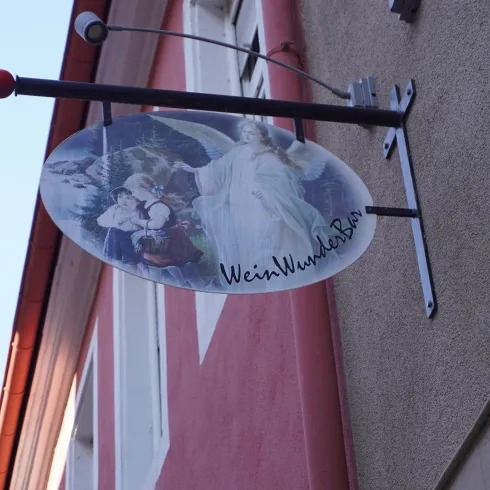 Ein hängendes Schild mit der Aufschrift "WeinWunderBar". Es zeigt eine künstlerische Darstellung von Personen und einer Landschaft.