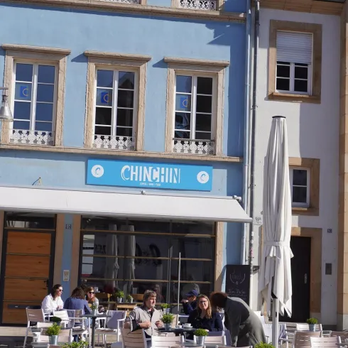 Ein charmantes Restaurant mit blauer Fassade und dem Namen „CHINCHIN“. Draußen sitzen Gäste an Tischen und genießen die Sonne.