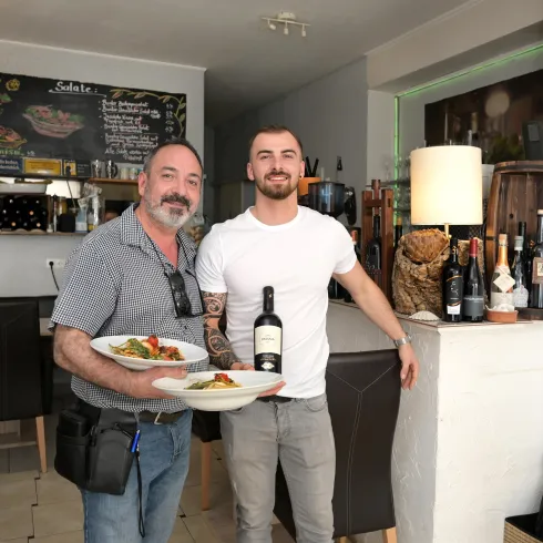 Zwei Männer stehen in einem Restaurant und halten Teller mit Speisen. Der eine Mann präsentiert eine Flasche Wein, während der andere lächelt.
