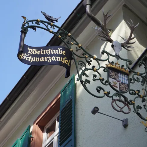 Ein künstlerisches Schild einer Weinstube mit dem Namen "Schwarzenfels". Das Schild zeigt detailreiche Verzierungen und Tiere.