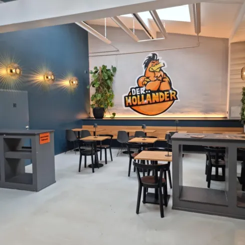 Ein modernes Restaurant mit minimalistischem Design. An der Wand befindet sich ein auffälliges Logo und es sind ein paar Pflanzen sowie Tische mit Stühlen zu sehen.