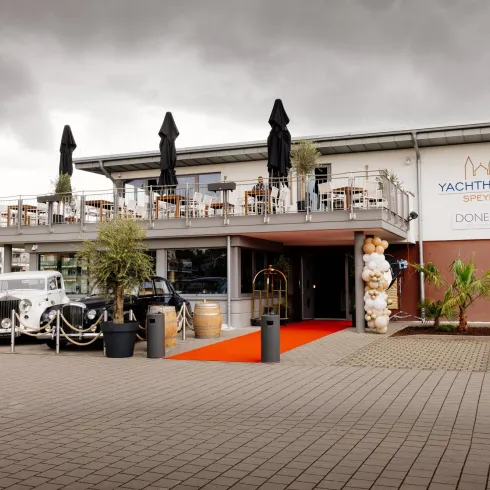 Ein modernes Restaurant mit einer Terrasse und Sonnenschirmen. Der Eingang ist mit einem roten Teppich und Pflanzen dekoriert.