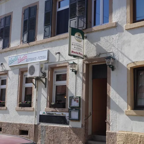 Ein Restaurant mit der Beschriftung "Pasta Pino" an der Fassadenwand. Die Außenansicht zeigt helle Wände und Fenster mit Fensterläden.