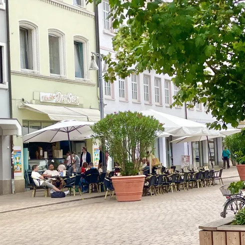 Eine belebte Straßencafé mit Tischen und Stühlen unter großen Sonnenschirmen. Menschen sitzen draußen und genießen die Atmosphäre.