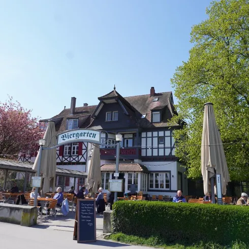 Ein malerisches Gasthaus mit einem Biergarten, umgeben von Bäumen und blühenden Pflanzen. Gäste genießen die Sonne an Tischen im Freien.