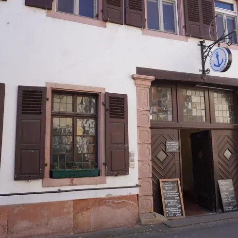 Ein historisches Gebäude mit einer einladenden Eingangstür und bunten Fenstern. An der Fassade hängen Schilder, die auf ein Restaurant hinweisen.