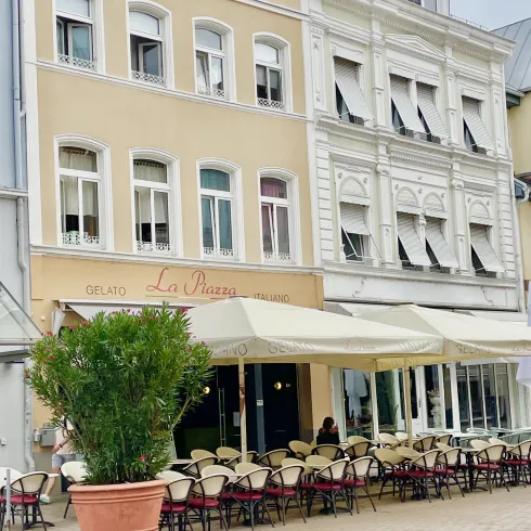 Ein einladendes Café mit Außenbereich und einem großen Topf mit Pflanzen. Die Fassade ist hell und die Fenster sind teils geöffnet.