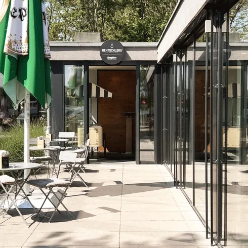 Ein modernes Café mit großen Glasfenstern und einer Terrasse. Es gibt Tische mit Stühlen und grüne Sonnenschirme.