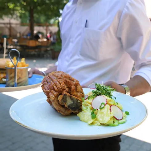 Ein Kellner trägt zwei Teller mit Essen. Auf einem Teller ist ein großer Burger und auf dem anderen Teller befindet sich ein gefüllter Kartoffelgericht mit frischem Salat.