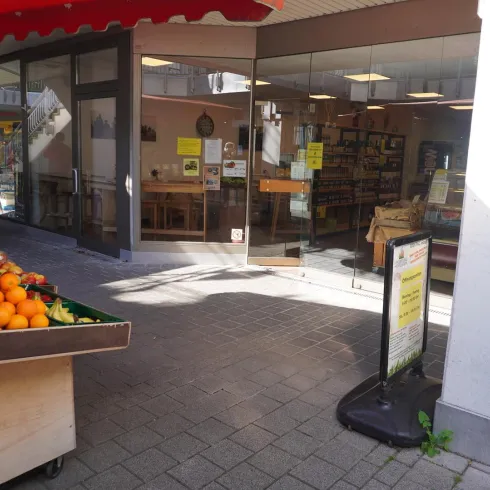 Ein Obststand mit einer Vielzahl frischer Früchte steht vor einem Laden. Im Hintergrund sind Tische und Regale sichtbar, die zum Einkaufen einladen.