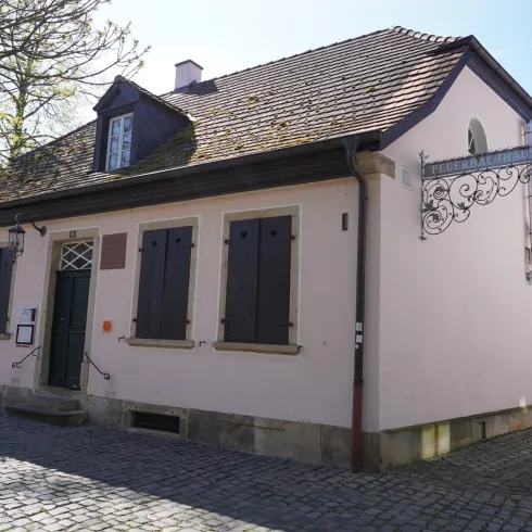 Ein charmantes, historisches Gebäude mit einem hellen Putz und geschlossenen Fensterläden. Der Pflasterweg vor dem Haus und die Bäume verleihen der Szenerie eine gemütliche Atmosphäre.