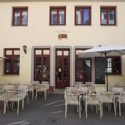 Ein gemütliches Restaurant mit einem hellen Eingang und vielen Stühlen im Freien. Ein Sonnenschirm sorgt für Schatten auf dem gepflasterten Platz.