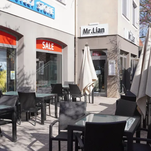 Ein gemütlicher Straßencafé-Bereich mit schwarzen Tischen und Stühlen. Markisen stehen zum Schutz vor Sonne und eine Sale-Anzeige ist sichtbar.