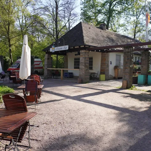 Ein gemütliches Café mit Holzstühlen und Tischen im Freien. Im Hintergrund sind Bäume und ein charmantes, kleines Gebäude zu sehen.