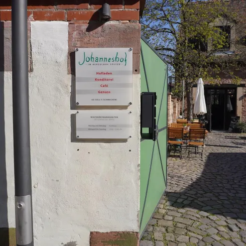 Ein Restaurant namens "Johanneshof" mit einem großen Schild. Im Hintergrund sind Tische und Stühle im Freien sowie ein paar Sonnenschirme zu sehen.