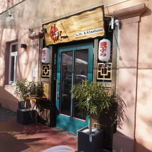 Ein asiatisches Restaurant mit einer bunten Eingangstür und dekorativen Laternen. Vor dem Restaurant stehen grüne Pflanzen.