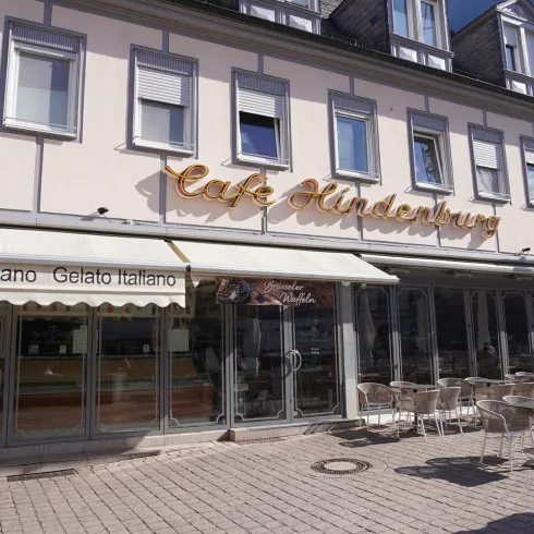 Ein charmantes Café mit dem Namen "Café Hindenburg". Die Fassade ist hell und einladend, mit Außenplätzen zum Sitzen.