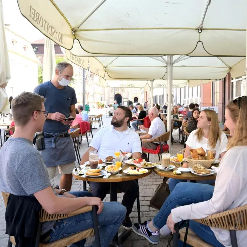 Eine Gruppe von Freunden sitzt in einem Café und genießt ihr Essen. Ein Kellner bedient sie unter einem großen Sonnenschirm.