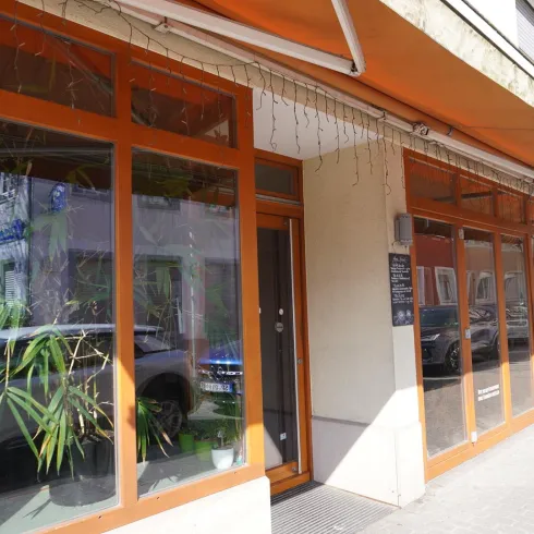 Ein modernes Geschäft mit großen Fenstern und oranger Markise. Im Inneren sind einige Pflanzen sichtbar.