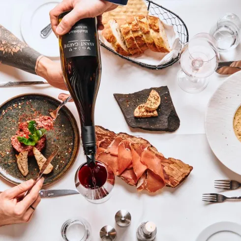 Ein festlich gedeckter Tisch mit verschiedenen Speisen, darunter Fleisch und eine cremige Pasta. Eine Person gießt Rotwein in ein Glas, während andere sich am Essen erfreuen.