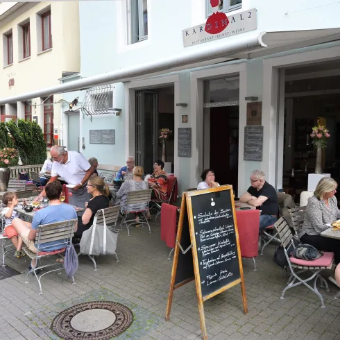 Ein belebtes Café mit außen sitzenden Gästen. Tische sind mit Stühlen umgeben, und eine Tafel zeigt die angebotenen Speisen.